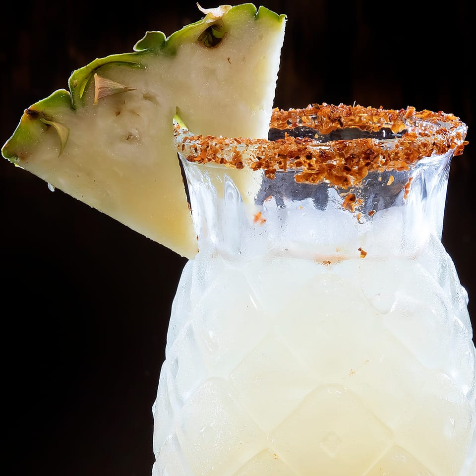A pineapple-shaped glass filled with a frosty drink, garnished with a pineapple slice and a chili-lime rim.