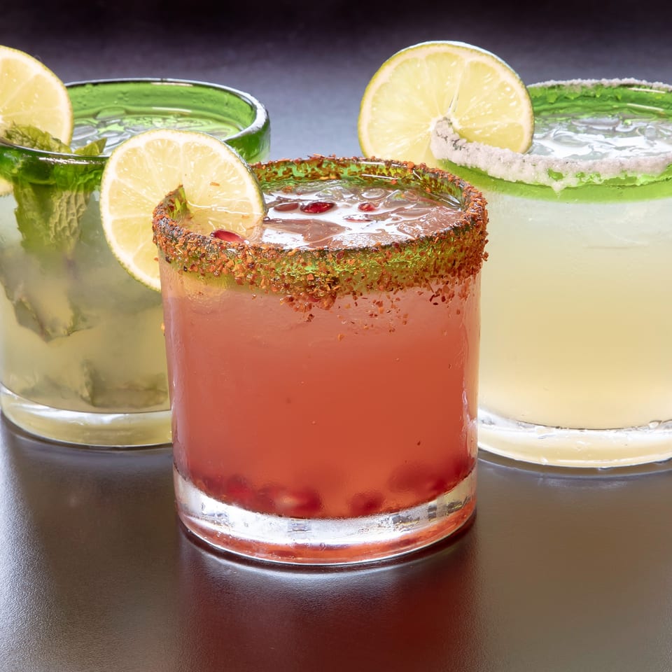 Three colorful Mexican cocktails: a mojito, a red drink with chili rim, and a margarita.