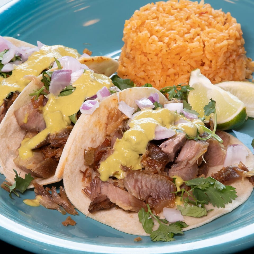 Three carnitas tacos with yellow sauce, red onions, cilantro, and a side of rice and lime wedges.