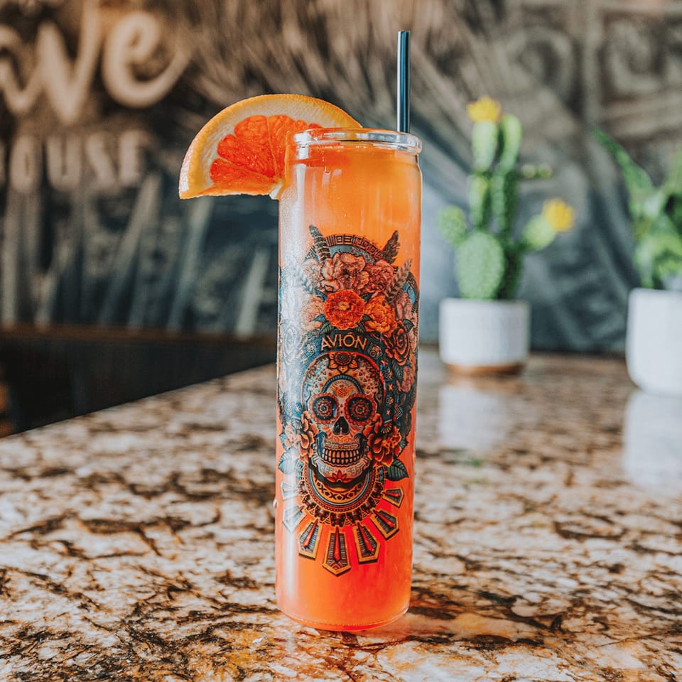A vibrant orange drink in a glass with a sugar skull design, garnished with a grapefruit slice.