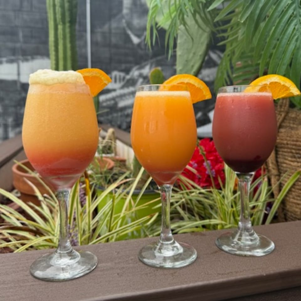 Three vibrant Los Jefes Grill cocktails, two orange and one red, garnished with orange slices.