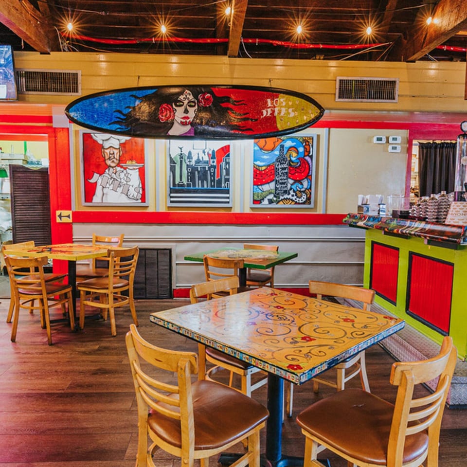 Colorful Los Jefes Grill interior with painted tables, wooden chairs, and vibrant Mexican-themed artwork on walls.