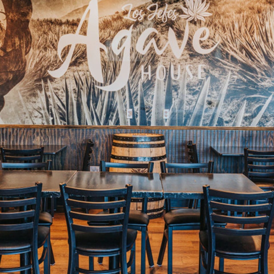Los Jefes Agave House restaurant interior with tables, chairs, and a barrel against a mural.