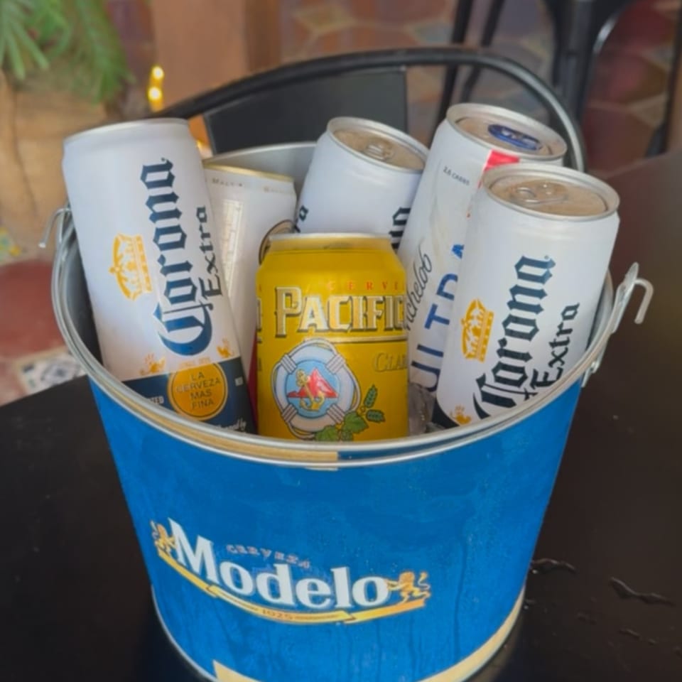 A blue Modelo branded bucket filled with various Mexican beer cans, including Corona Extra and Pacifico.