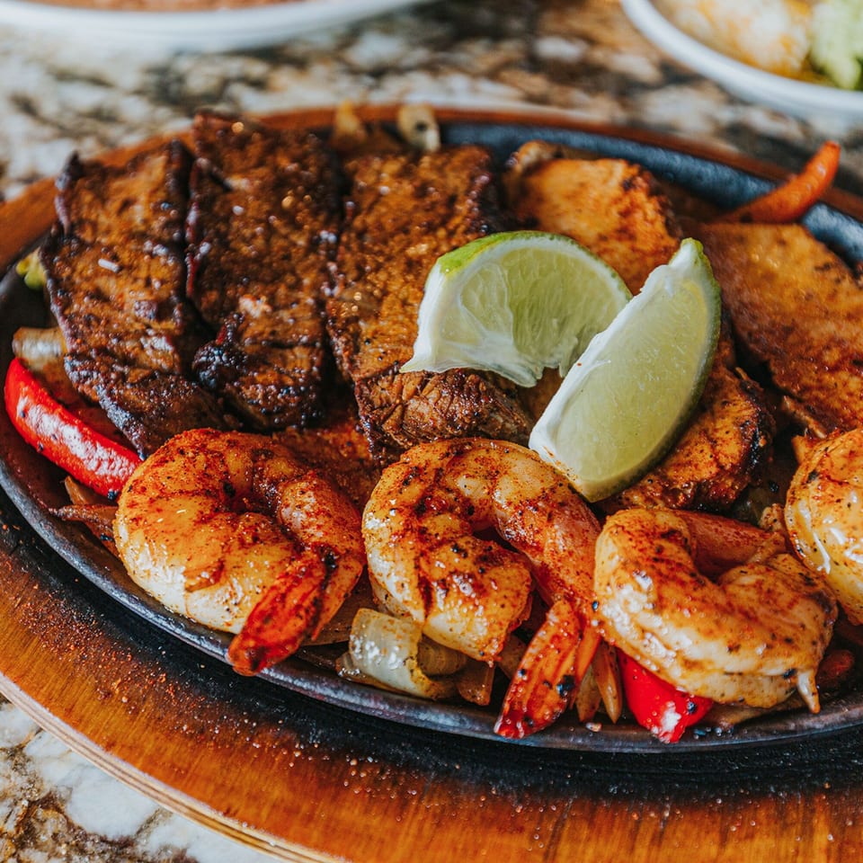 Sizzling fajitas with grilled steak, shrimp, peppers, onions, and lime wedges on a hot plate.